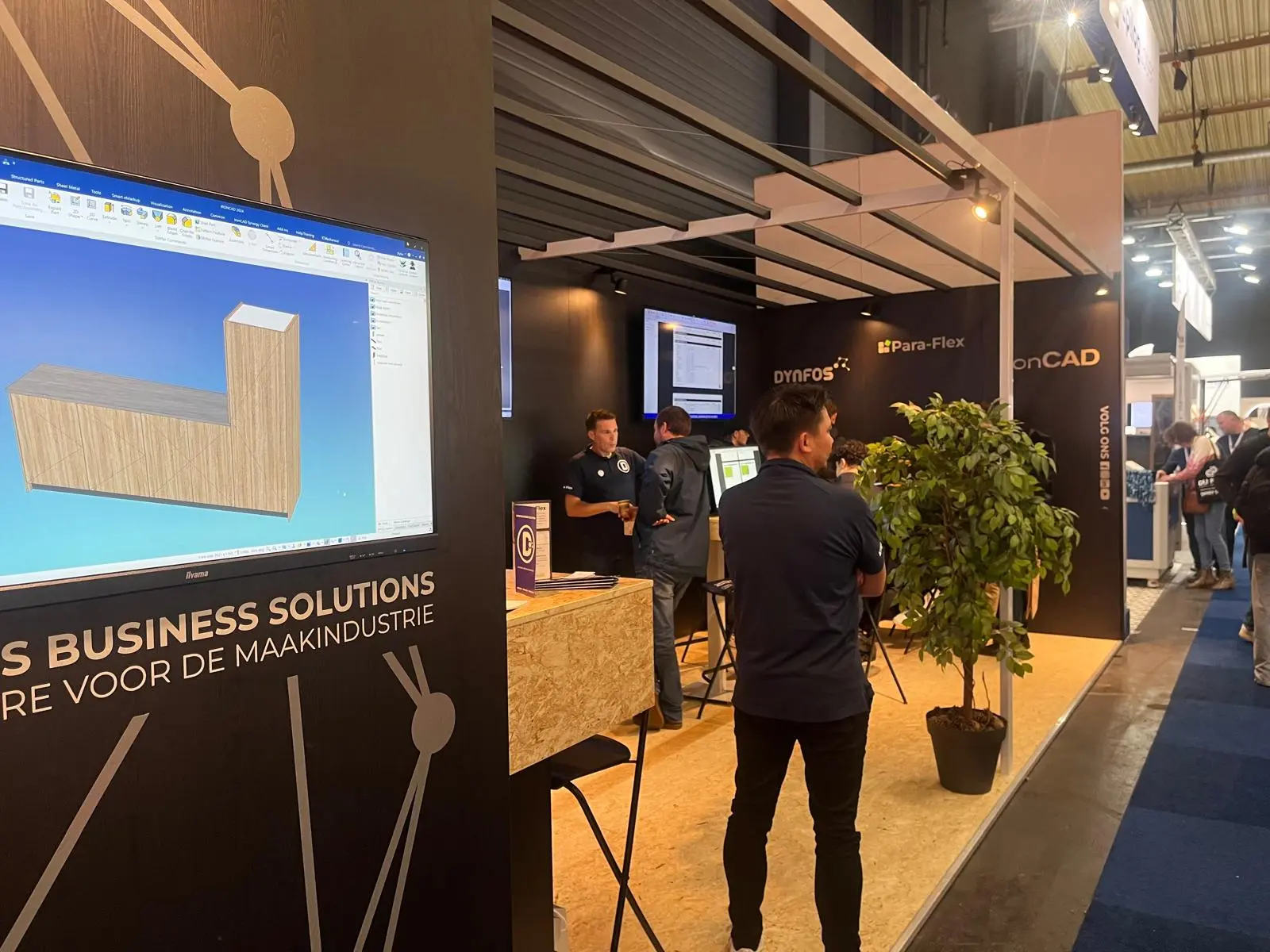 Beursstand met scherm waarop 3D-ontwerp zichtbaar is, bezoekers in gesprek en demonstratie.