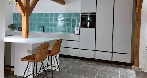 “Moderne designkeuken van Design en Meer met witte greeploze kasten, turquoise tegelwand, houten balken en kookeiland met twee barkrukken.”