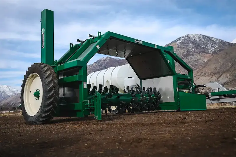 Compostomzetter van Midwest Bio-Systems in actie op landbouwgrond, met grote wielen en roterende elementen voor het mengen van organisch materiaal, tegen een bergachtig landschap.