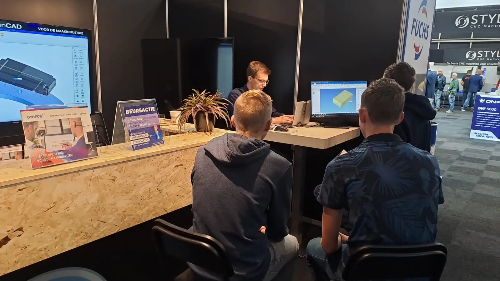 Bezoekers bekijken 3D-ontwerpdemonstratie aan een beursstand, met schermen en uitleg door een medewerker.
