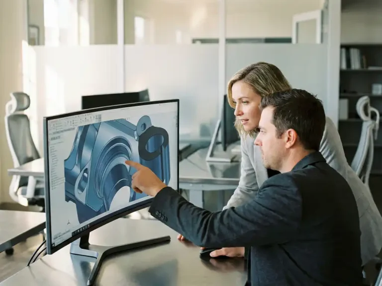 Designer wijst gebogen mechanische onderdelen aan in 3D CAD-model op monitor, nieuwsgierige klant kijkt mee in modern kantoor.